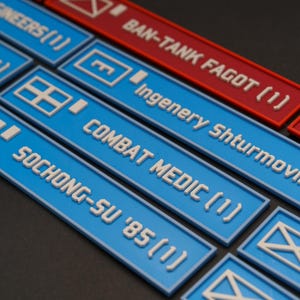 Könnte beinhalten: Nahaufnahme von mehreren rechteckigen Spielsteinen. Die meisten sind blau mit weißem Text, darunter "SOCHONG-SU '85 (1)" und "COMBAT MEDIC (1)". Einer ist rot mit "BAN-TANK FAGOT (1)". Jedes Teil hat ein weißes Symbol.