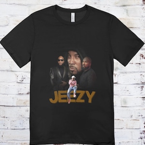 May include: Black t-shirt featuring a graphic of three men and the word "JEEZY" in gold lettering. The graphic includes a person wearing a white hat and pink shirt. The shirt is made of a soft material.
