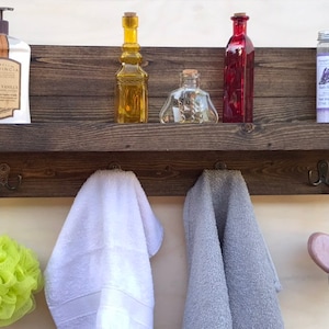 May include: A rustic wooden shelf with hooks, featuring a white towel, a gray towel, a green bath puff, and various bottles and jars. The shelf has a dark brown stain and is made of reclaimed wood.