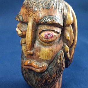 Op de afbeelding: Een handgesneden houten sculptuur van een hoofd met een baard, een prominente wenkbrauw en twee kleine hoorns aan de bovenkant. Het gezicht heeft wijde, uitpuilende ogen met rood en wit geschilderde details, donker gesneden haar en een textuur.