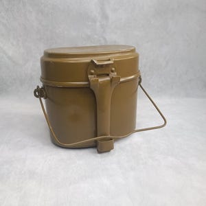 May include: An olive drab metal mess kit with an oval shape, featuring a secure locking latch on the front and a thin, hinged wire handle attached to the sides. The container has a smooth, solid finish and a flat lid that fits snugly over the base.