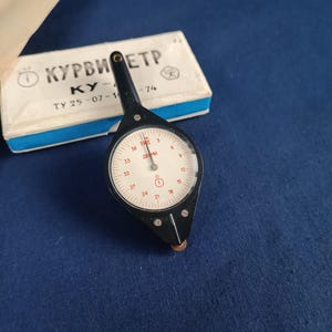 May include: A black and silver vintage instrument with a circular dial and a needle. The dial has numbers and markings. A white box with Cyrillic text is in the background. The instrument is likely used for measuring or navigation.