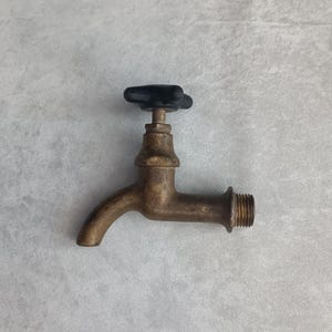 May include: A brass water faucet featuring a curved spout and a black, four-pointed star-shaped handle. The faucet body has a weathered, metallic finish, and the threaded inlet pipe is visible on the right side for secure installation.