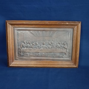 May include: A framed relief sculpture of the Last Supper. The artwork is in a rectangular wooden frame. The relief shows a detailed depiction of the biblical scene, with figures seated around a table. The background is a detailed architectural setting.