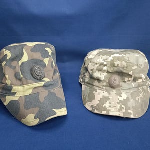 Pode incluir: Duas boinas de estilo militar com padrões de camuflagem. Uma boina apresenta um design de camuflagem castanho, verde e bege, enquanto a outra tem um padrão de camuflagem digital em tons de cinza e bege. Ambas as boinas têm um emblema circular.