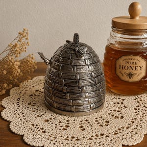 May include: A silver-toned metal honey pot shaped like a traditional skep beehive with a small bee figurine on the lid, positioned next to a glass jar of honey with a wooden lid and a label that reads Pure Honey with a bee illustration.