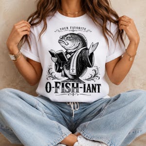 以下が含まれることがあります： 眼鏡とタキシードを着用し、開いた本を持っている魚の黒いグラフィックイラストが描かれた白いTシャツ。魚の上には「YOUR FAVORITE」、下には太字の装飾フォントで「O-FISH-IANT」というテキストが配置されています。