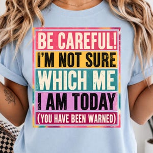 May include: A light blue t-shirt featuring a rectangular graphic with a distressed texture. The text reads: BE CAREFUL! I'M NOT SURE WHICH ME I AM TODAY (YOU HAVE BEEN WARNED). The design uses horizontal bands of pink, yellow, teal, and purple.