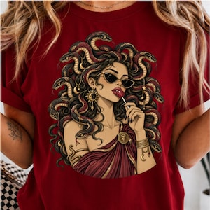 Peut inclure: Un t-shirt rouge foncé avec une illustration graphique représentant une silhouette aux cheveux en forme de serpents, portant des lunettes de soleil œil-de-chat et tenant une sucette rouge. Elle porte des bijoux dorés, dont une boucle d'oreille soleil.