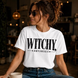 May include: A white short-sleeved t-shirt featuring the black text WITCHY, OBVIOUSLY. printed on the chest. The text is styled with a distressed, textured appearance and is underlined with a line flanked by two small four-pointed star icons.