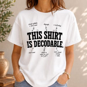 May include: A white short-sleeved t-shirt featuring the black text THIS SHIRT IS DECODABLE. Arrows point to specific words with labels including closed syllable, digraph, r-controlled, short vowel, silent-e makes the vowel say its name, and Latin-based ending = able.