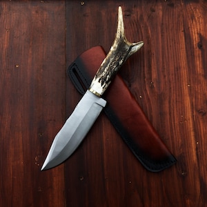 Puede incluir: Un cuchillo de caza con una hoja de acero pulido y un mango de asta. El cuchillo descansa sobre una funda de cuero marr&oacute;n con costuras negras. El cuchillo y la funda se colocan sobre un fondo de madera oscura.