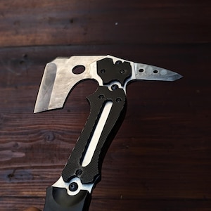 May include: A silver and black tactical tomahawk axe with a sharp blade and a pointed end. The handle is black with a textured grip, and the axe head has a hole. The axe is designed for outdoor use and survival.