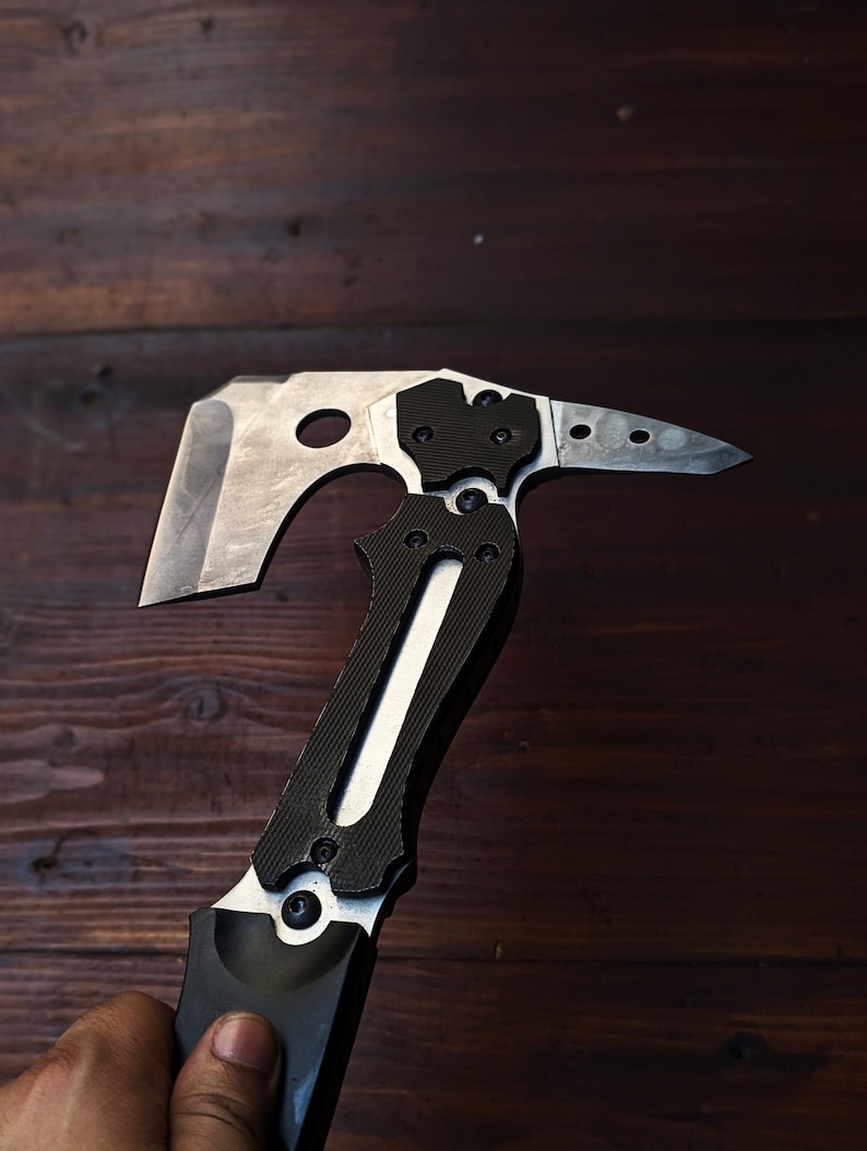 May include: A silver and black tactical tomahawk axe with a sharp blade and a pointed end. The handle is black with a textured grip, and the axe head has a hole. The axe is designed for outdoor use and survival.