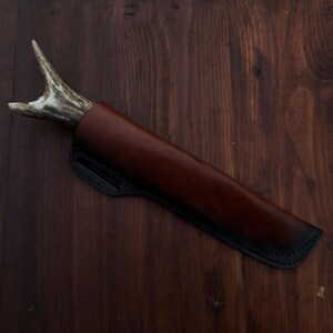 Puede incluir: Un cuchillo artesanal con mango de asta, enfundado en una funda de cuero marr&oacute;n con costuras negras. El cuchillo se presenta sobre un fondo de madera oscura, mostrando la artesan&iacute;a y la est&eacute;tica r&uacute;stica.