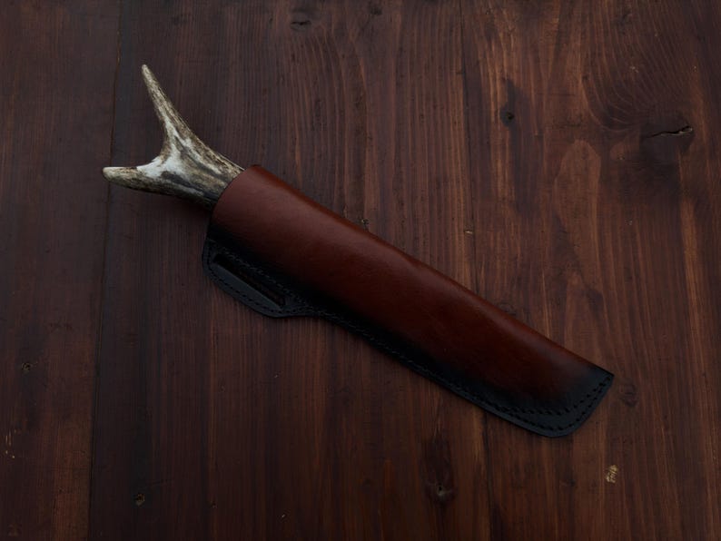 Puede incluir: Un cuchillo artesanal con mango de asta, enfundado en una funda de cuero marr&oacute;n con costuras negras. El cuchillo se presenta sobre un fondo de madera oscura, mostrando la artesan&iacute;a y la est&eacute;tica r&uacute;stica.