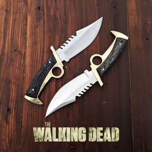 May include: Two silver and black knives with gold accents and serrated blades on a brown wooden background. The text "THE WALKING DEAD" is printed below the knives.
