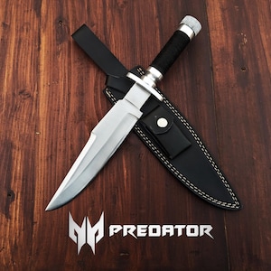 May include: A silver hunting knife with a black leather sheath. The knife has a black handle and a silver blade. The sheath has white stitching and the word "PREDATOR" is printed on the wooden surface.