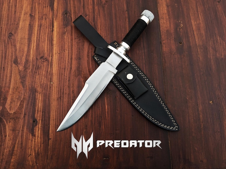 May include: A silver hunting knife with a black leather sheath. The knife has a black handle and a silver blade. The sheath has white stitching and the word "PREDATOR" is printed on the wooden surface.