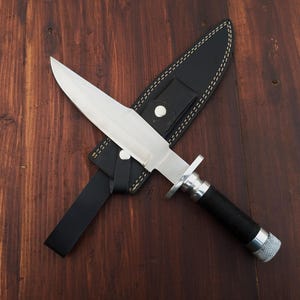 May include: A silver knife with a black handle and a leather sheath. The knife is lying on a wooden surface.
