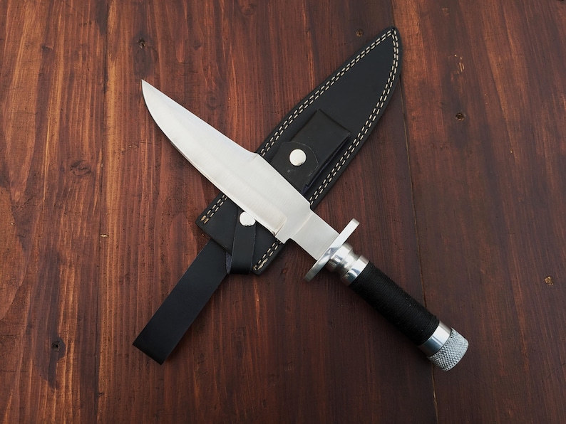 May include: A silver knife with a black handle and a leather sheath. The knife is lying on a wooden surface.