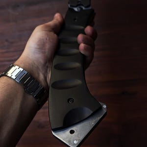 May include: A black tactical axe with a silver blade is held in a hand. The axe has a textured handle and a silver blade. The background is a dark wood surface.