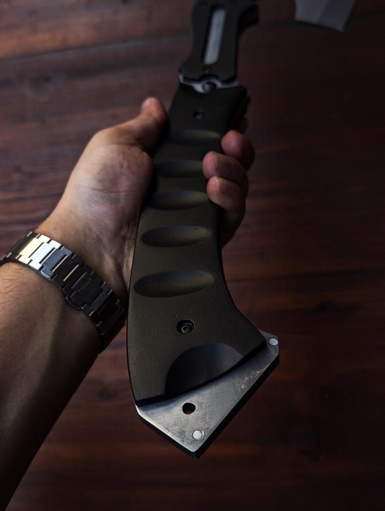 May include: A black tactical axe with a silver blade is held in a hand. The axe has a textured handle and a silver blade. The background is a dark wood surface.