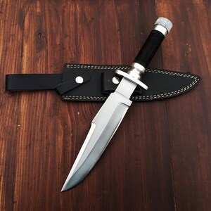 May include: A silver-colored hunting knife with a black handle and a leather sheath. The knife is lying on a wooden surface.