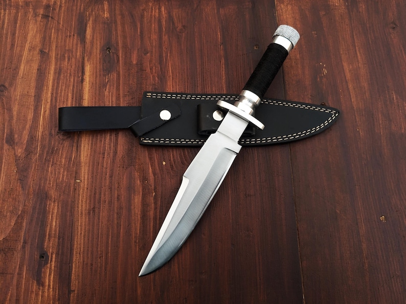 May include: A silver-colored hunting knife with a black handle and a leather sheath. The knife is lying on a wooden surface.