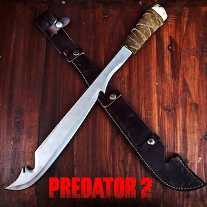 Könnte beinhalten: Ein großes, gebogenes Messer mit silberner Oberfläche, einem umwickelten Griff und einer Lederscheide. Der Text "PREDATOR 2" ist unten rot dargestellt. Das Messer liegt über der Scheide auf einer Holzoberfläche.