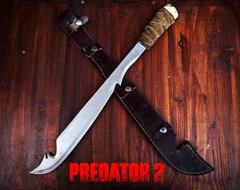 May include: A large, curved blade knife with a silver finish, a wrapped handle, and a leather sheath. The text "PREDATOR 2" is displayed in red at the bottom. The knife is laid across the sheath on a wooden surface.