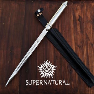Può includere: Un pugnale argentato e nero con un fodero in pelle. Il pugnale ha una lama lunga e appuntita e un manico argentato. Il fodero è nero. L'immagine include anche il testo "SUPER NATURAL" e un simbolo solare.