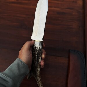 Puede incluir: Un cuchillo de caza grande, hecho a mano, con una hoja de acero pulido y un mango de asta de ciervo. El cuchillo tiene una virola de lat&oacute;n y una funda de cuero. La hoja mide aproximadamente 20 cm de largo.