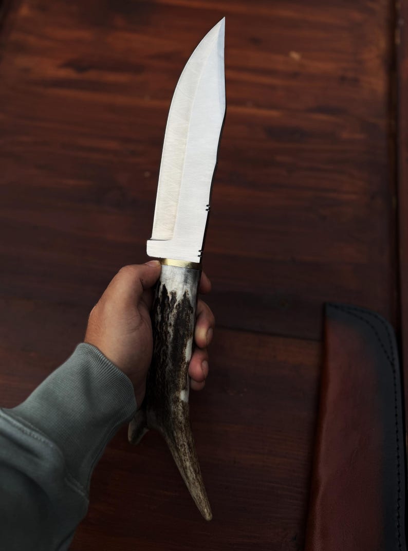 Puede incluir: Un cuchillo de caza grande, hecho a mano, con una hoja de acero pulido y un mango de asta de ciervo. El cuchillo tiene una virola de lat&oacute;n y una funda de cuero. La hoja mide aproximadamente 20 cm de largo.