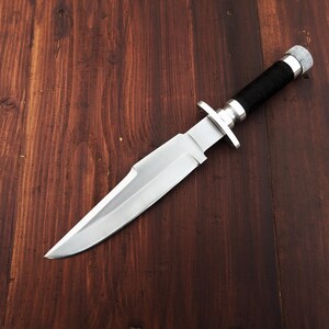May include: A silver hunting knife with a black handle on a wooden surface.