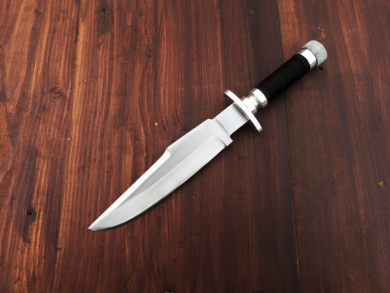 May include: A silver hunting knife with a black handle on a wooden surface.