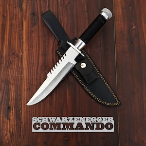 May include: A silver knife with a black handle and a serrated blade. The knife is in a black leather sheath with yellow stitching. The knife is on a brown wooden surface. The text "SCHWARZENEGGER COMMANDO" is on the surface.