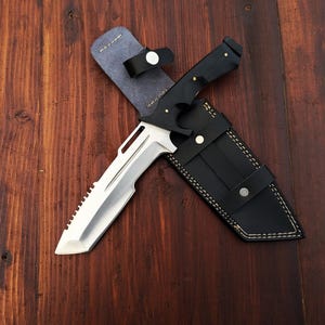 Può includere: Un coltello argentato con manico nero e lama seghettata è mostrato in un fodero di cuoio nero con cuciture bianche. Il coltello è su una superficie di legno.