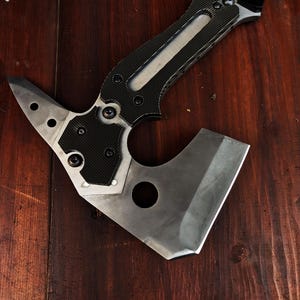 May include: A tactical axe with a silver blade and a black and silver handle. The axe head features a pointed end with three holes and a curved blade. The handle has a textured grip for secure handling.