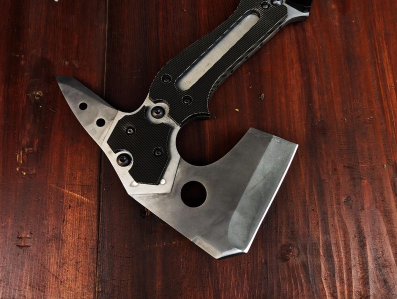 May include: A tactical axe with a silver blade and a black and silver handle. The axe head features a pointed end with three holes and a curved blade. The handle has a textured grip for secure handling.