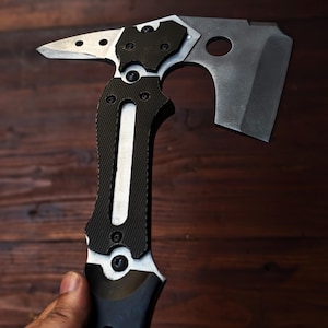 May include: A tactical axe with a black and silver design. The axe features a hammer, a sharp blade, and a textured handle for grip. The axe is held in a hand against a dark wood background.