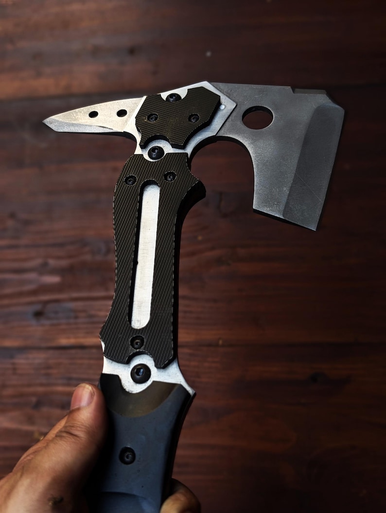 May include: A tactical axe with a black and silver design. The axe features a hammer, a sharp blade, and a textured handle for grip. The axe is held in a hand against a dark wood background.