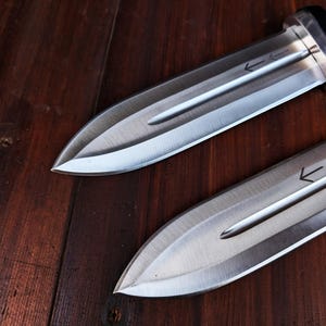 Puede incluir: Dos dagas de doble filo con hojas de metal plateado que presentan un canal central y s&iacute;mbolos de flechas grabados. La daga superior tiene un mango negro, mientras que la daga inferior cuenta con una guarda de tono lat&oacute;n.