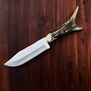 Puede incluir: Un cuchillo de caza artesanal con una hoja de acero pulido y un mango de asta de ciervo. El cuchillo se encuentra sobre un fondo de madera oscura, mostrando su dise&ntilde;o r&uacute;stico y su filo.
