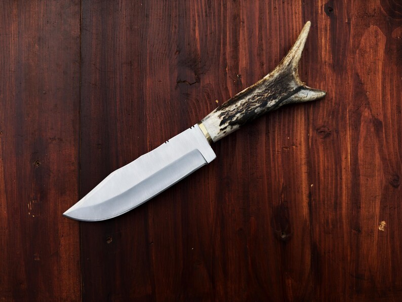 Puede incluir: Un cuchillo de caza artesanal con una hoja de acero pulido y un mango de asta de ciervo. El cuchillo se encuentra sobre un fondo de madera oscura, mostrando su dise&ntilde;o r&uacute;stico y su filo.