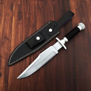May include: A silver-colored hunting knife with a black handle and a leather sheath. The knife is lying on a wooden surface.