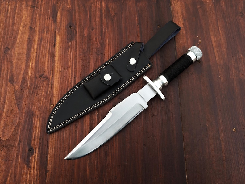 May include: A silver-colored hunting knife with a black handle and a leather sheath. The knife is lying on a wooden surface.