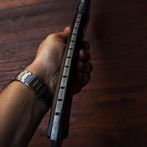 May include: A dark-handled knife with a silver blade and rectangular cutouts along the spine. The knife is held against a dark wooden background. A silver watch is visible on the wrist.