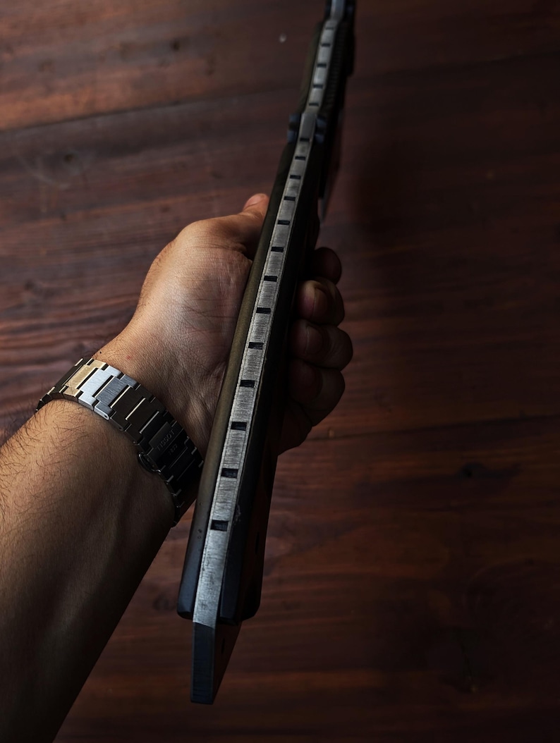 May include: A dark-handled knife with a silver blade and rectangular cutouts along the spine. The knife is held against a dark wooden background. A silver watch is visible on the wrist.