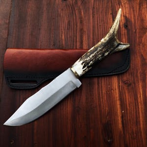 Puede incluir: Cuchillo de caza artesanal con una hoja de acero pulido y un mango de asta. El cuchillo descansa sobre una funda de cuero marr&oacute;n oscuro, sobre un fondo de madera. La hoja mide unos 20 cm.